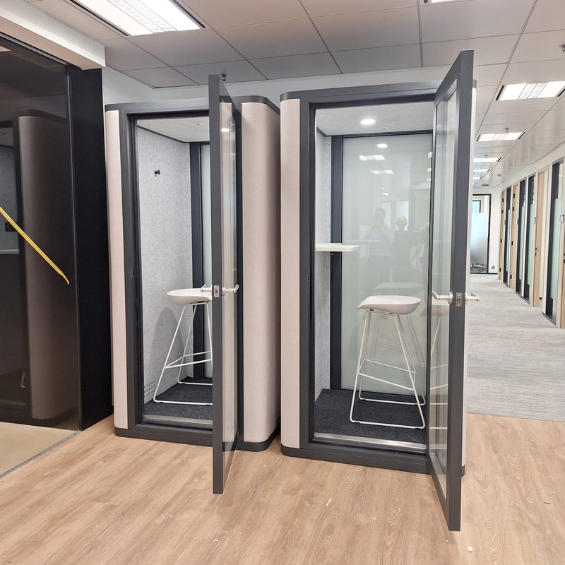 Two office phone booths with stools inside in a modern office setting.